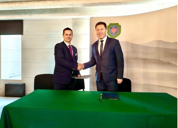 Dwaj eleganccy mężczyźni w garniturach ściskają sobie dłonie nad zielonym stołem, na tle godła i g&oacute;rskiego krajobrazu.